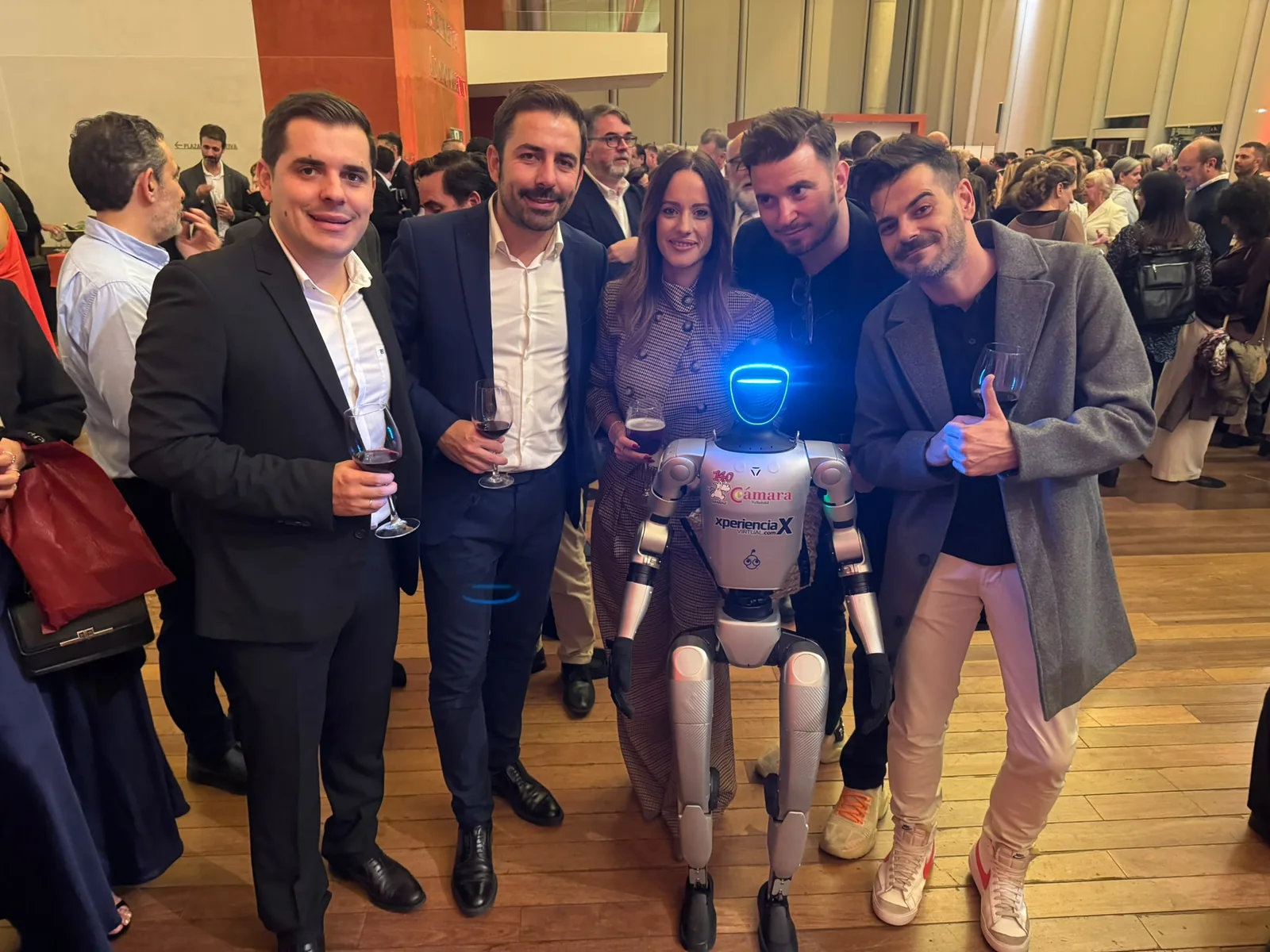 El robot Unitree G1, con el logo del 140 aniversario de la Cámara y el de Xperiencia Virtual en el pecho, posa con invitados durante el cóctel